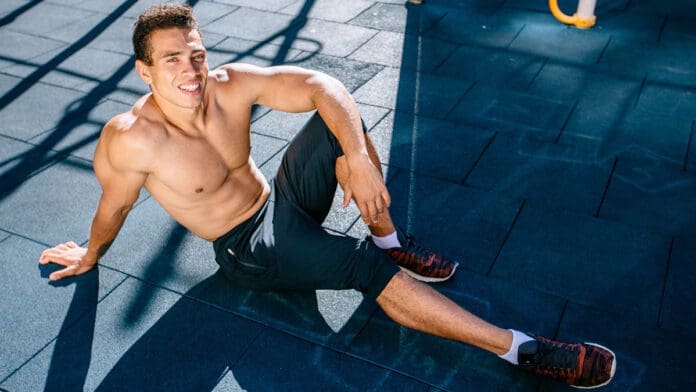 Sports. Smiling Man With Fit Body In Yellow Sportswear Resting on Gray Grunge After Running, Jogging Outdoors. Handsome Healthy Male Runner Taking Break After Fitness Workout And Exercising.